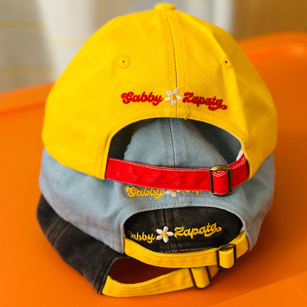 Three baseball caps stacked together on an orange surface. The top cap is bright yellow with red 'Gabby Zapata' embroidery and a red back strap.