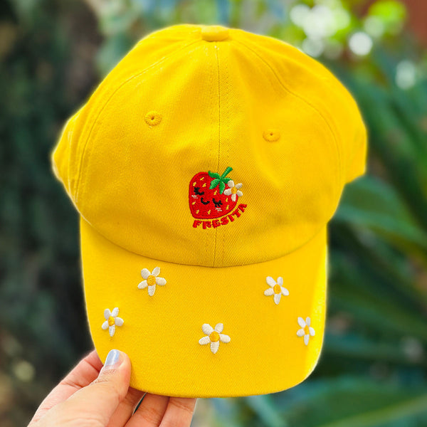 A bright yellow baseball cap held by a hand, featuring an embroidered red strawberry with a smiling face and the word 'FRESITA' on the front, with small white flower appliqués on the brim, against a blurred green garden background.