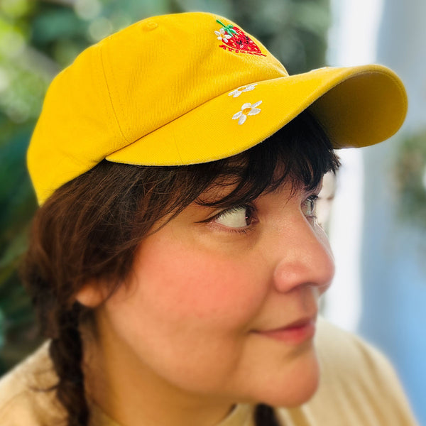 A close-up of a person wearing a bright yellow baseball cap with embroidered details, including a strawberry motif and white flowers, against a softly blurred outdoor background.