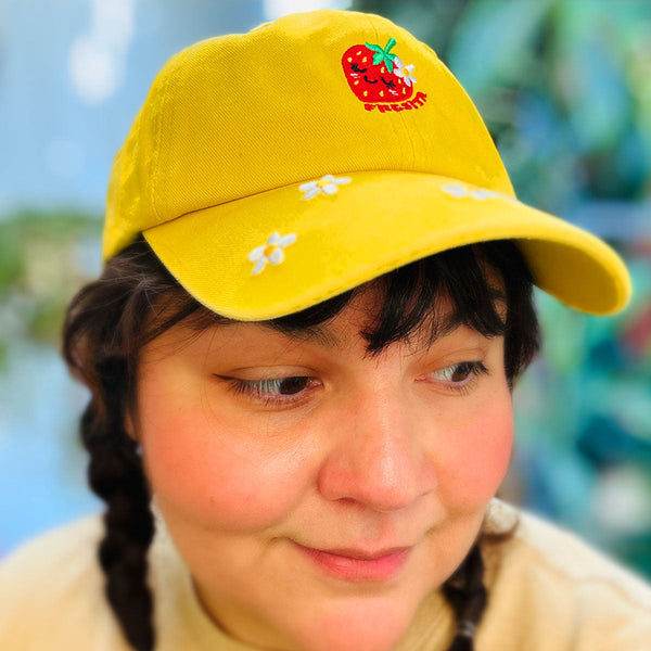 A close-up of a person wearing a bright yellow baseball cap with embroidered white flowers and a red strawberry on the front, text reading "FRESITA" photographed against a softly blurred outdoor background.