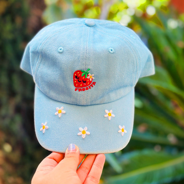A light blue baseball cap held by a hand, featuring a red embroidered strawberry with the word 'FRESITA' on the front and small white daisy embellishments on the brim, set against a blurred green garden background.
