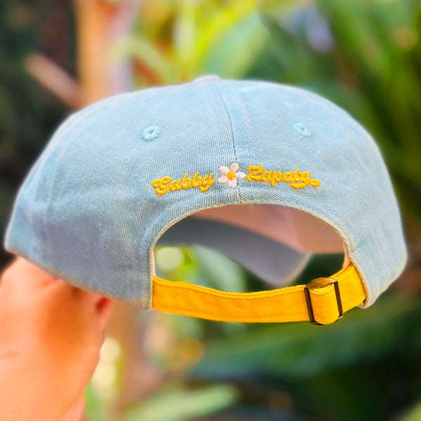 A light blue denim baseball cap held by a hand, featuring yellow embroidered text reading 'Gabby Zapata' with a small white flower in the center. The cap has a yellow adjustable strap and is photographed against a blurred green outdoor background.