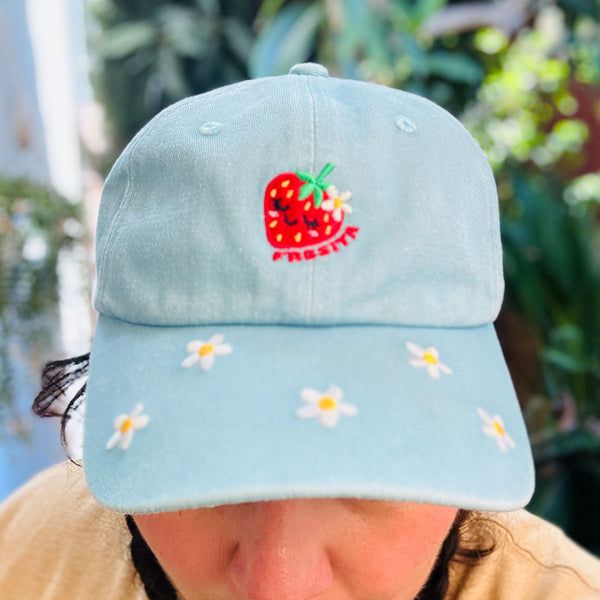 A light blue cotton baseball cap with embroidered white daisies on the brim and a red strawberry patch on the front. The cap is being worn by a person, with a blurred green garden background. Reads "FRESITA"