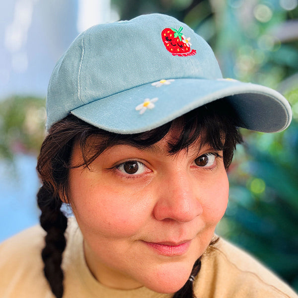 A close-up portrait of a woman wearing a light blue baseball cap with embroidered white daisies and a red strawberry patch on the front, reads "FRESITA" set against a blurred green and blue outdoor background.