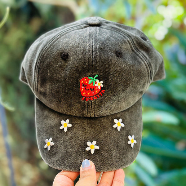 A dark gray washed baseball cap held by a hand, featuring an embroidered red strawberry with a white flower and the word 'FRESITA' on the front, and five white daisy flowers embroidered on the brim, against a blurred green garden background.