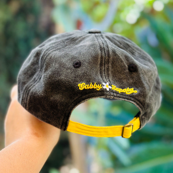 A dark gray washed baseball cap held by a hand, featuring yellow embroidered 'GABBY ZAPATA' text and a yellow adjustable strap against a blurred green outdoor background.