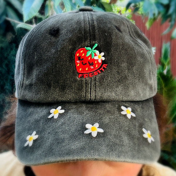 A close-up of a person wearing a dark gray washed baseball cap with embroidered white daisies on the brim and a red strawberry patch on the front. Reads "FRESITA" The background shows blurred green foliage and a red fence.