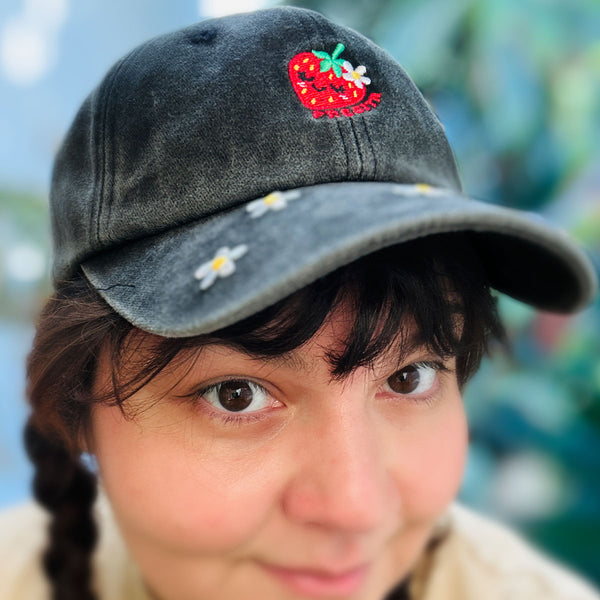 A close-up of a person wearing a dark gray washed baseball cap with a red strawberry patch and small white daisy embroidery on the brim, against a blurred green background. Reads "FRESITA" embroidered